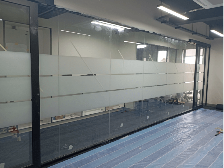 Glass partition with frosted horizontal bands in an empty office under renovation; blue floor protection visible beneath.